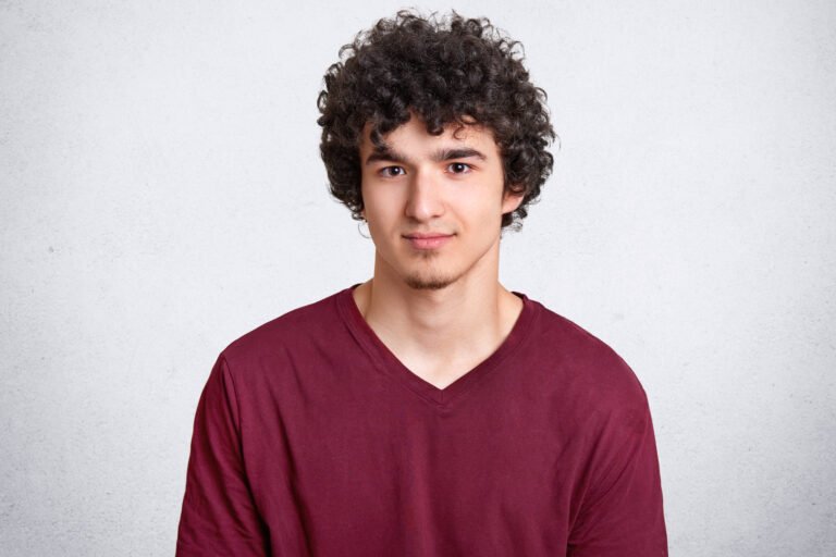 Headshot of attractive curly youngster looks seriously at camera, has appealing look, spends free time with friends, dressed in casual outfit, isolated over white concrete wall